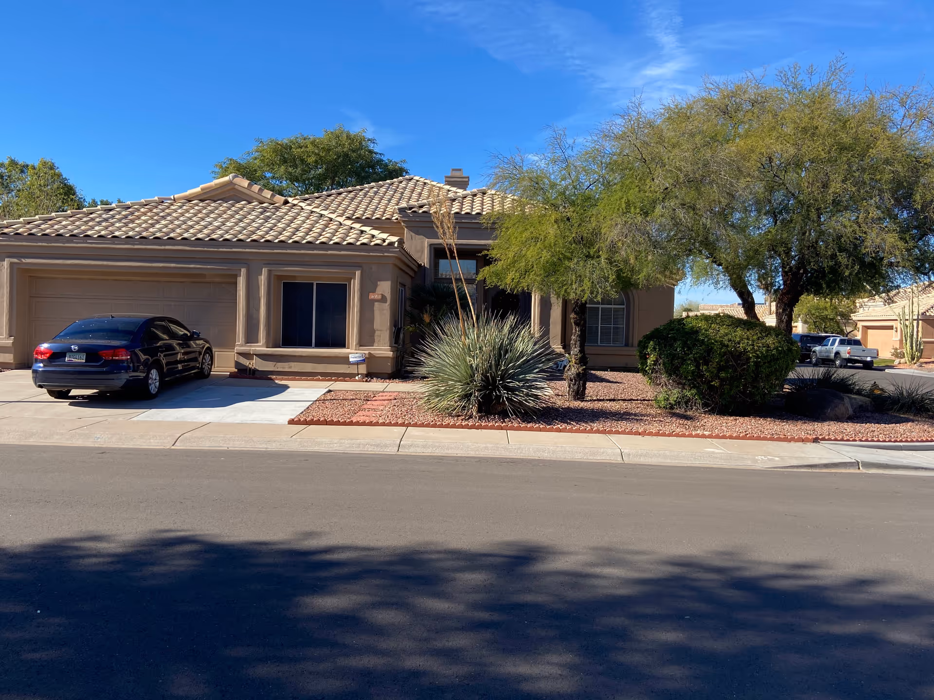 Front exterior view of a single-story residential building with a tiled roof, a two-car garage, and a driveway with a parked black car. The front yard features desert landscaping with rocks, bushes, and trees under a clear blue sky.