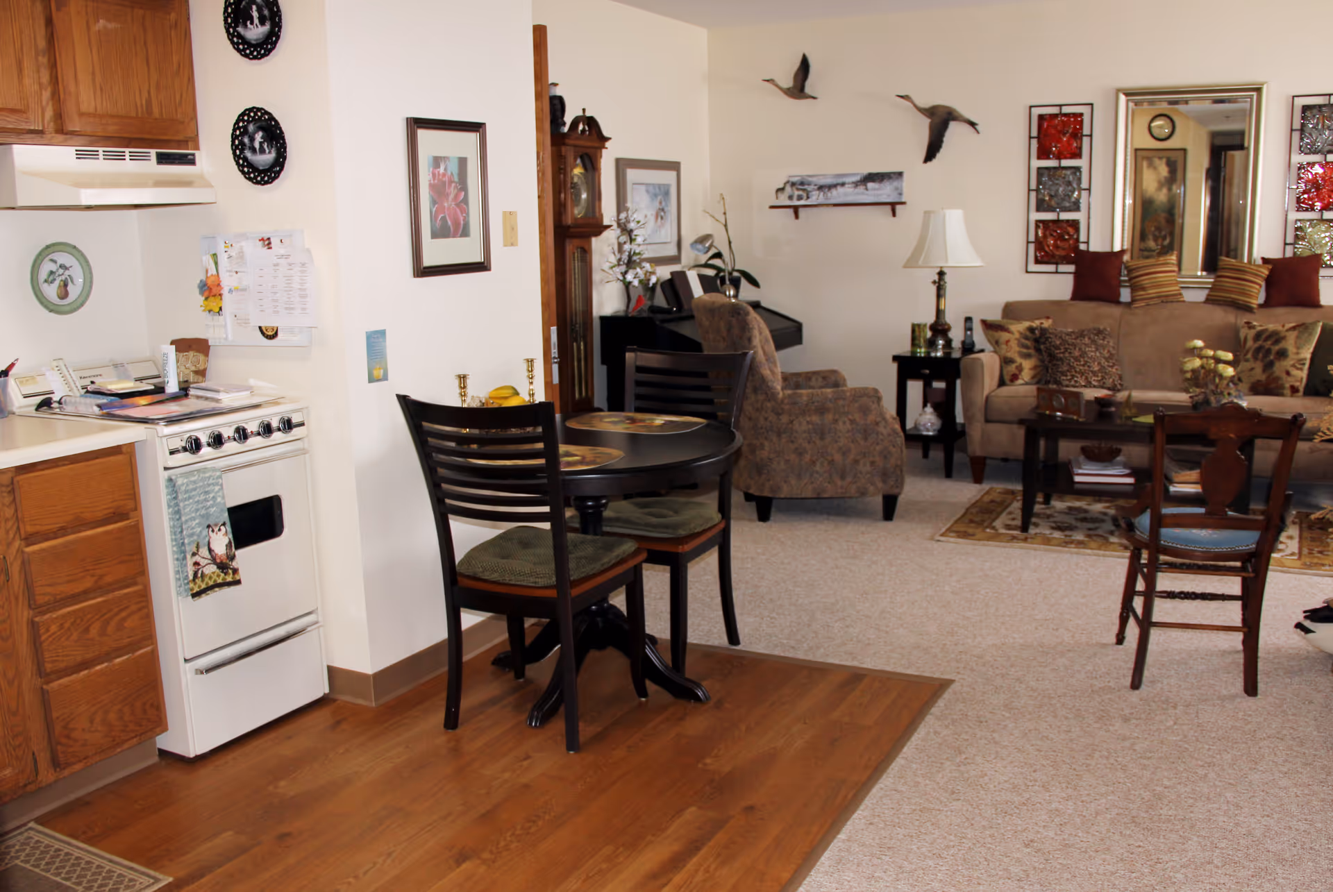 Open-plan living area showing a small kitchen and round dining table opening onto a cozy living room with a sofa, armchairs, and decorative wall art.