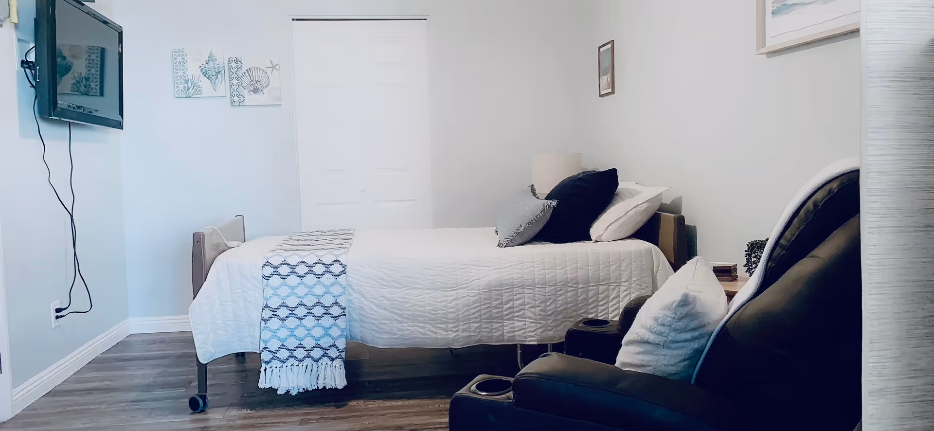 A neatly arranged bedroom in a senior living facility with a single bed covered in white bedding and a patterned throw blanket. There are several pillows on the bed. A black recliner chair with a white pillow is positioned near the bed. The room has light-colored walls with minimal decorations, including two framed pictures on the wall and a wall-mounted TV with visible cords. The floor is wooden, and there is a closed white closet door in the background.