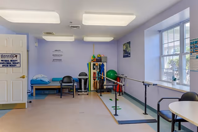 A physical therapy room with light purple walls and ceiling lights. The room contains parallel bars for walking exercises, a green exercise ball, various therapy equipment on shelves, a bench with pillows, and chairs. A window on the right side lets in natural light. A door on the left has a sign that reads 'Genesis Physical Therapy Occupational Therapy Speech Therapy.'