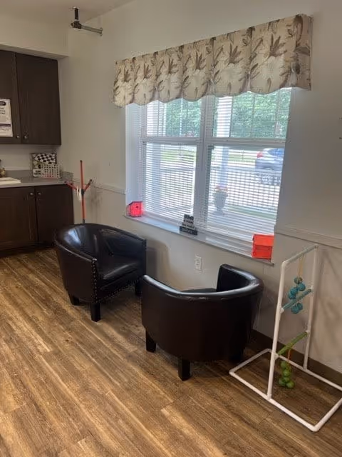 Two black leather armchairs positioned near a window with blinds and a floral valance. The room has wood flooring and a white wall with a small white rack holding green and blue balls. A kitchen counter with dark cabinets is partially visible on the left side.