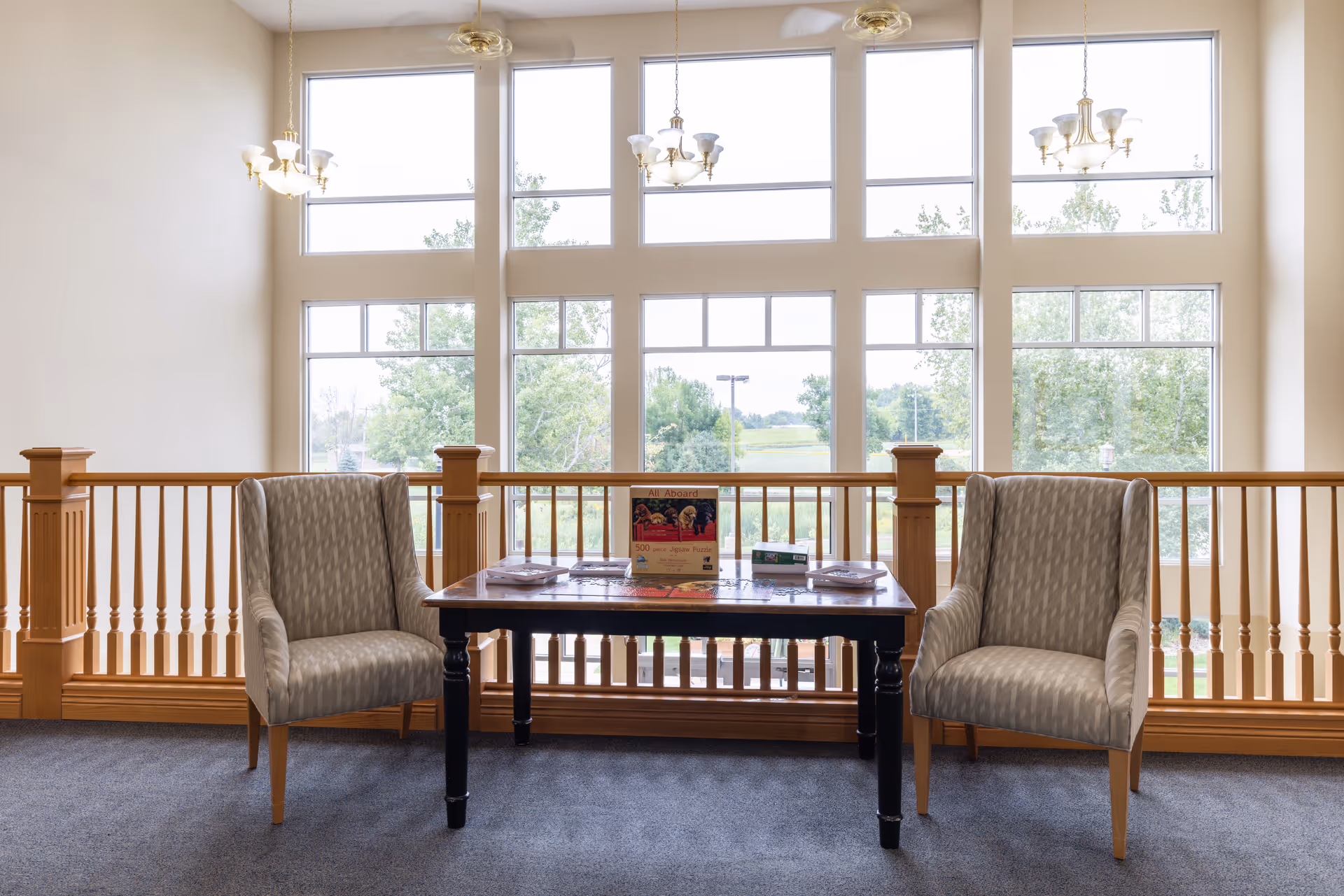A cozy sitting area with two patterned armchairs facing a wooden table that holds a puzzle box and some puzzle pieces. The area is well-lit by large windows behind the table, showing a view of trees and greenery outside. The space features wooden railing and carpeted flooring, with three chandeliers hanging from the ceiling.