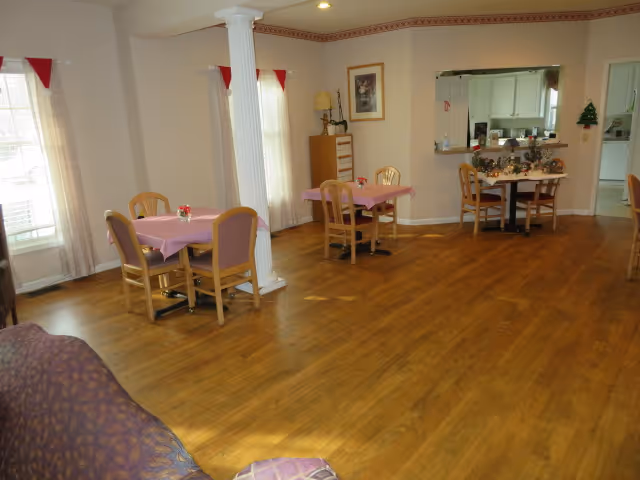 A spacious room with wooden flooring featuring several small dining tables covered with pink tablecloths and surrounded by wooden chairs. The room has large windows with curtains, a white decorative column, and a pass-through window to a kitchen area decorated with holiday garlands. A purple patterned couch is partially visible in the foreground.