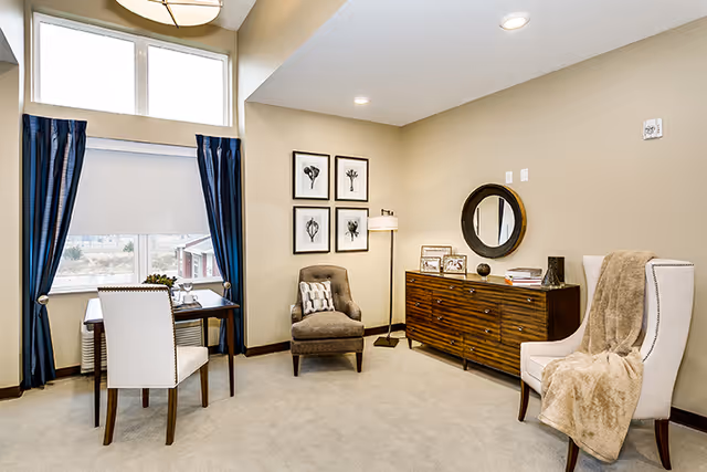 A cozy living room area with beige walls and carpet. The room features a wooden sideboard with decorative items and a round mirror above it, a beige armchair with a brown throw blanket, a gray cushioned chair with a patterned pillow, a floor lamp, and a small table with two white chairs near a window with blue curtains and a white roller shade.