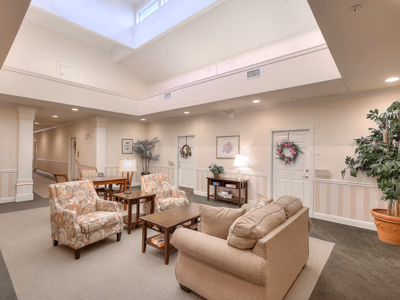 A bright and spacious common area in a senior living facility featuring a beige sofa, two patterned armchairs, a wooden coffee table, and side tables with lamps. The room has high ceilings with skylights, light-colored walls with wainscoting, decorative wreaths on doors, framed artwork, and potted plants. A hallway extends from the seating area.