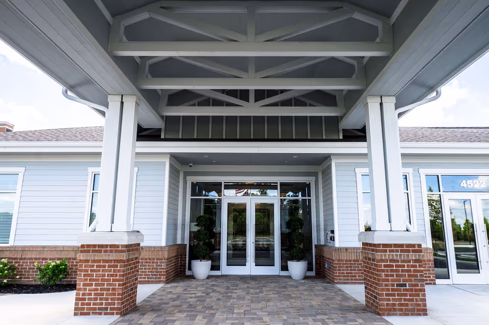 Senior living facility entrance with glass doors, covered walkway, brick columns, and potted plants.