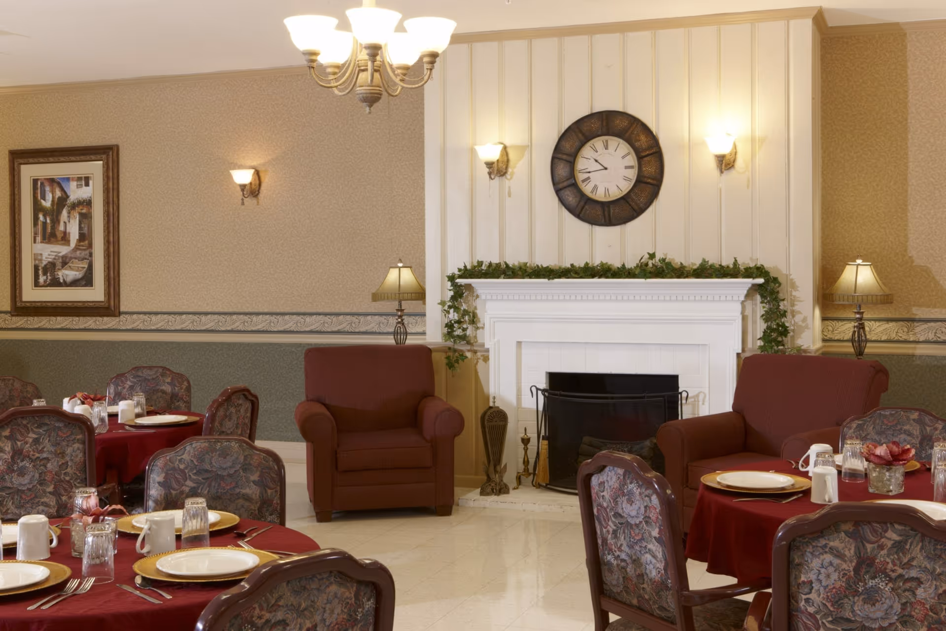 A cozy dining area with round tables covered in red tablecloths set with plates, glasses, and silverware. There are floral upholstered chairs around the tables. In the background, there are two red armchairs flanking a white fireplace decorated with green garland and a large round clock above it. The walls have patterned wallpaper and there are several lamps and wall sconces providing warm lighting.
