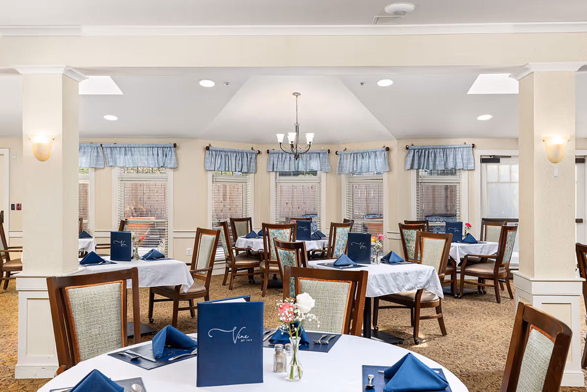 A bright dining room with multiple tables covered in white tablecloths and set with blue napkins, menus, and small flower vases. The room has several windows with blue valances, beige walls, and carpeted floors. Wooden chairs with cushioned seats surround the tables.