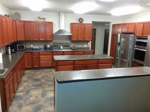 A spacious kitchen with wooden cabinets, granite countertops, a stainless steel refrigerator, double wall ovens, a microwave, toaster, coffee maker, and a stovetop with a stainless steel range hood. The floor has a tiled pattern and the walls are painted white.