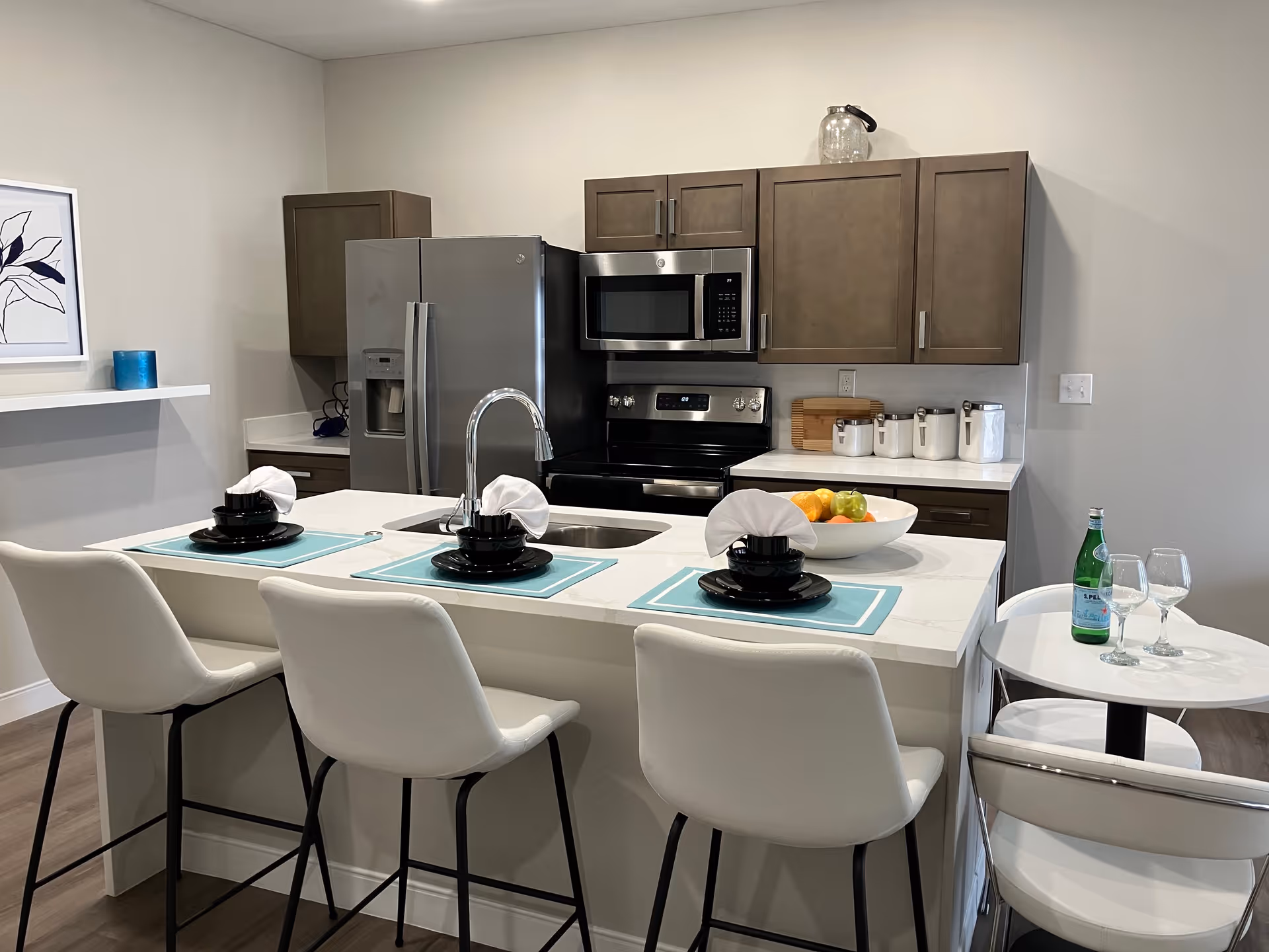 Modern kitchen with a white island countertop featuring a sink and three place settings with black dishes and white napkins on blue placemats. Behind the island, there is a stainless steel refrigerator, microwave, and stove with brown cabinets above and below the countertops. A bowl of fruit and several white canisters are on the counter. To the right, a small round table with two chairs holds a bottle of sparkling water and two wine glasses. The walls are light-colored and there is a framed abstract artwork on the left wall.