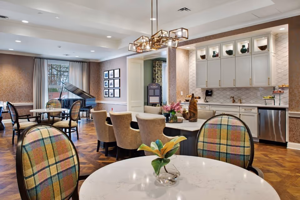 A well-lit interior common area featuring a seating arrangement with round tables and upholstered chairs, a grand piano near a window with curtains, and a kitchen area with white cabinets, a countertop, and modern lighting fixtures.