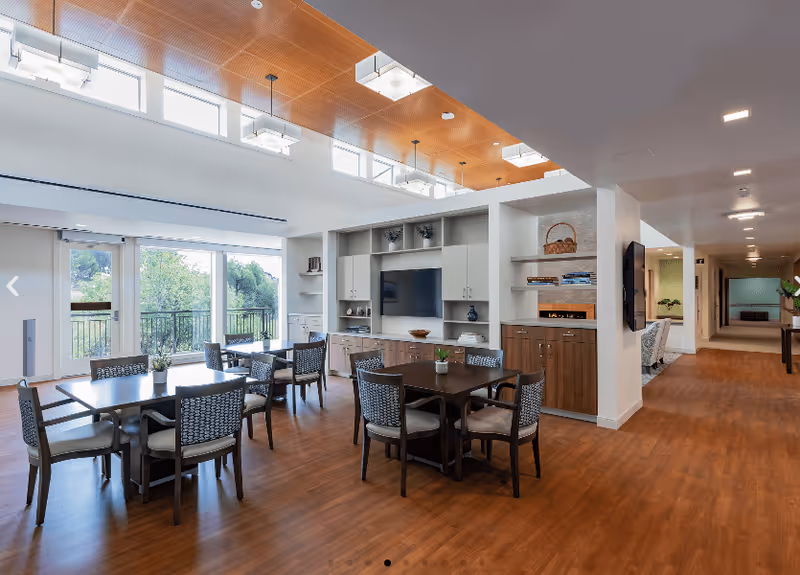 Bright communal dining and lounge area with multiple tables and chairs, built-in cabinetry and TV, and large windows opening to a balcony.