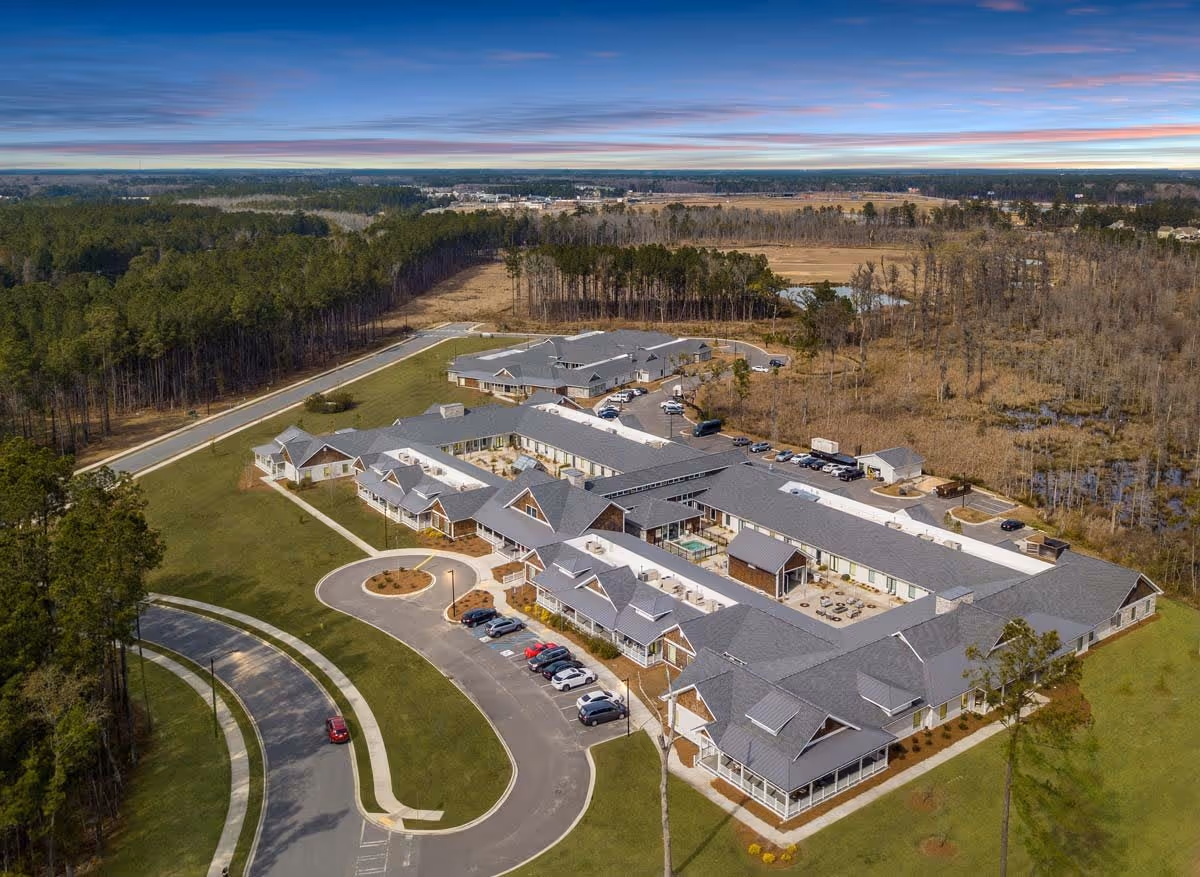 Aerial view of Legacy Savannah Quarters senior living facility surrounded by trees and open land, showing multiple connected buildings with gray roofs, parking areas with cars, and a curved driveway leading to the entrance.