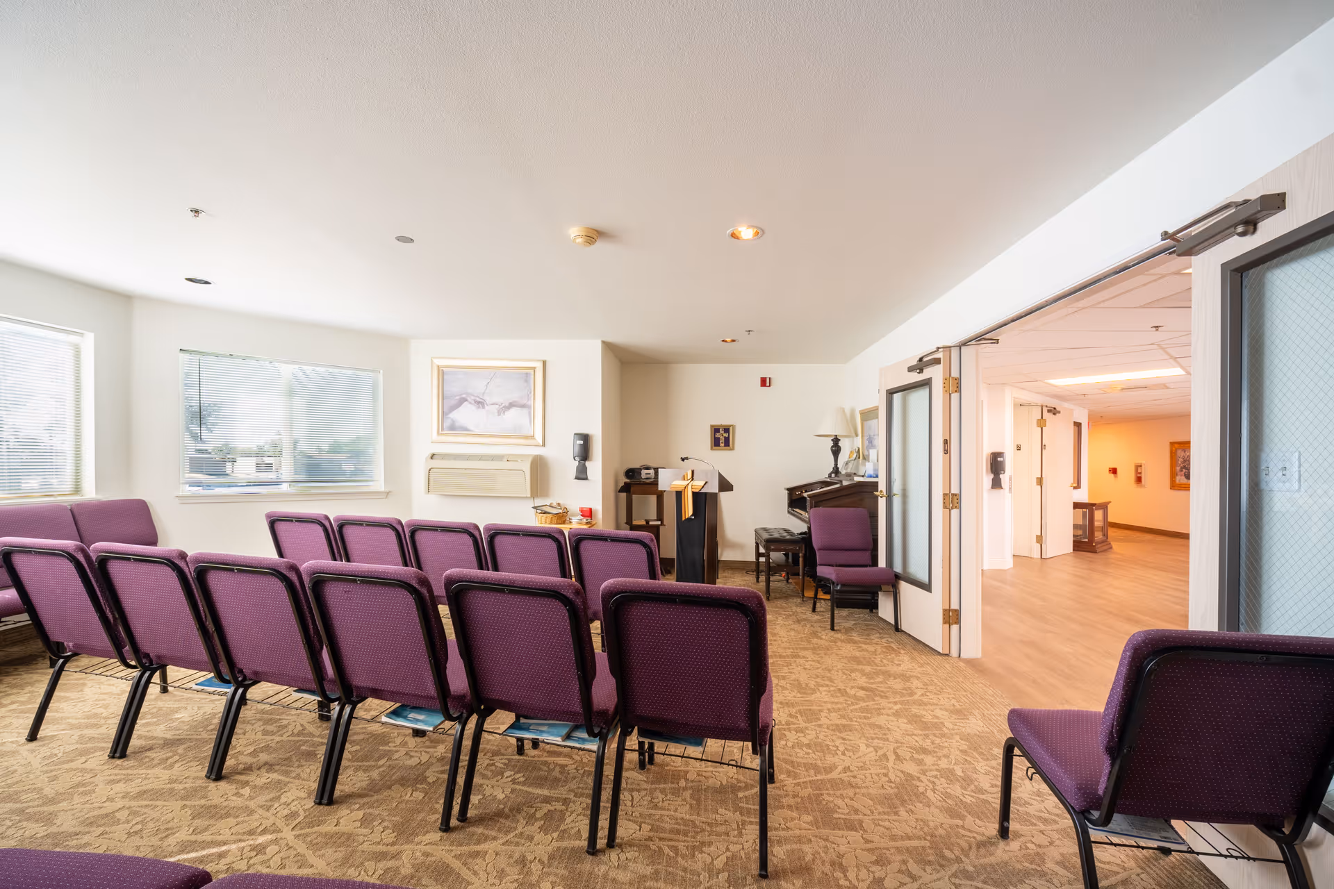 A small meeting or chapel room with rows of purple chairs facing a podium and a piano. The room has beige carpet, white walls, two windows with blinds, and a framed picture above an air conditioning unit. Double doors on the right side open to a hallway with wood flooring and light-colored walls.