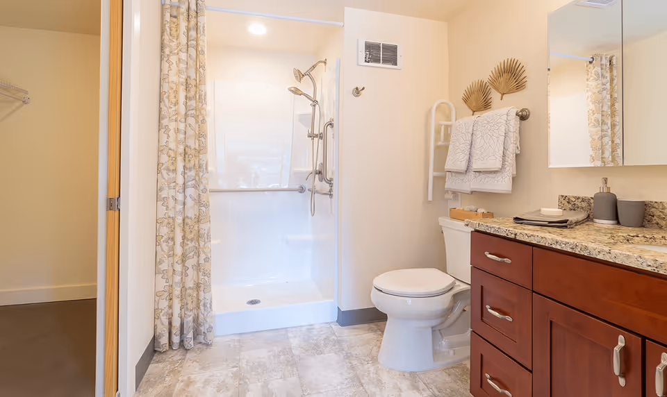 A clean and well-lit bathroom featuring a white toilet, a shower with a floral patterned curtain, and a wooden vanity with a granite countertop. Two decorative palm leaf wall hangings and neatly folded towels are visible above the toilet.
