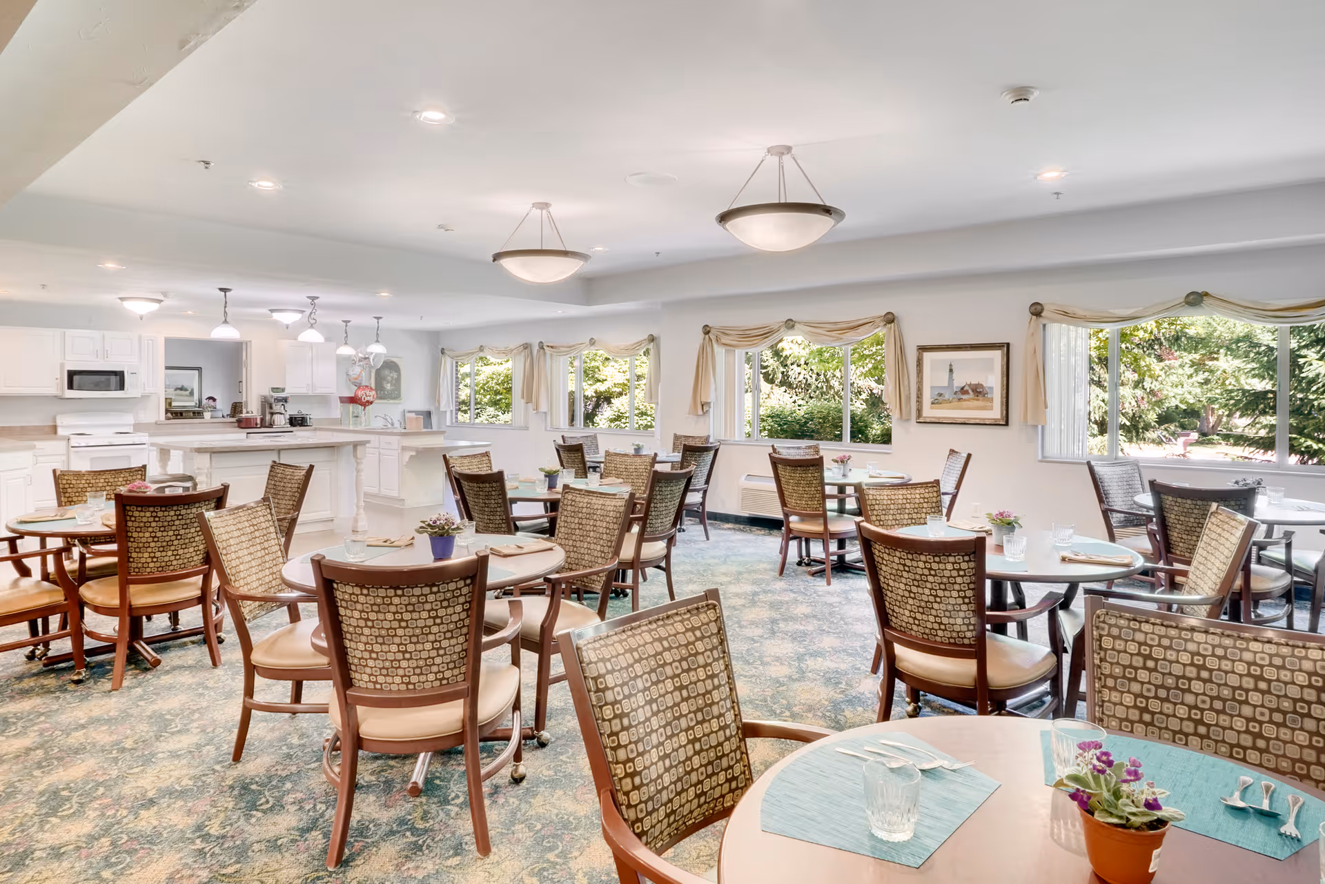 A bright dining room with multiple round tables and chairs arranged neatly. Each table has placemats, utensils, glasses, and small potted plants. Large windows with light curtains allow natural light to fill the room, and a kitchen area with white cabinets and appliances is visible in the background.