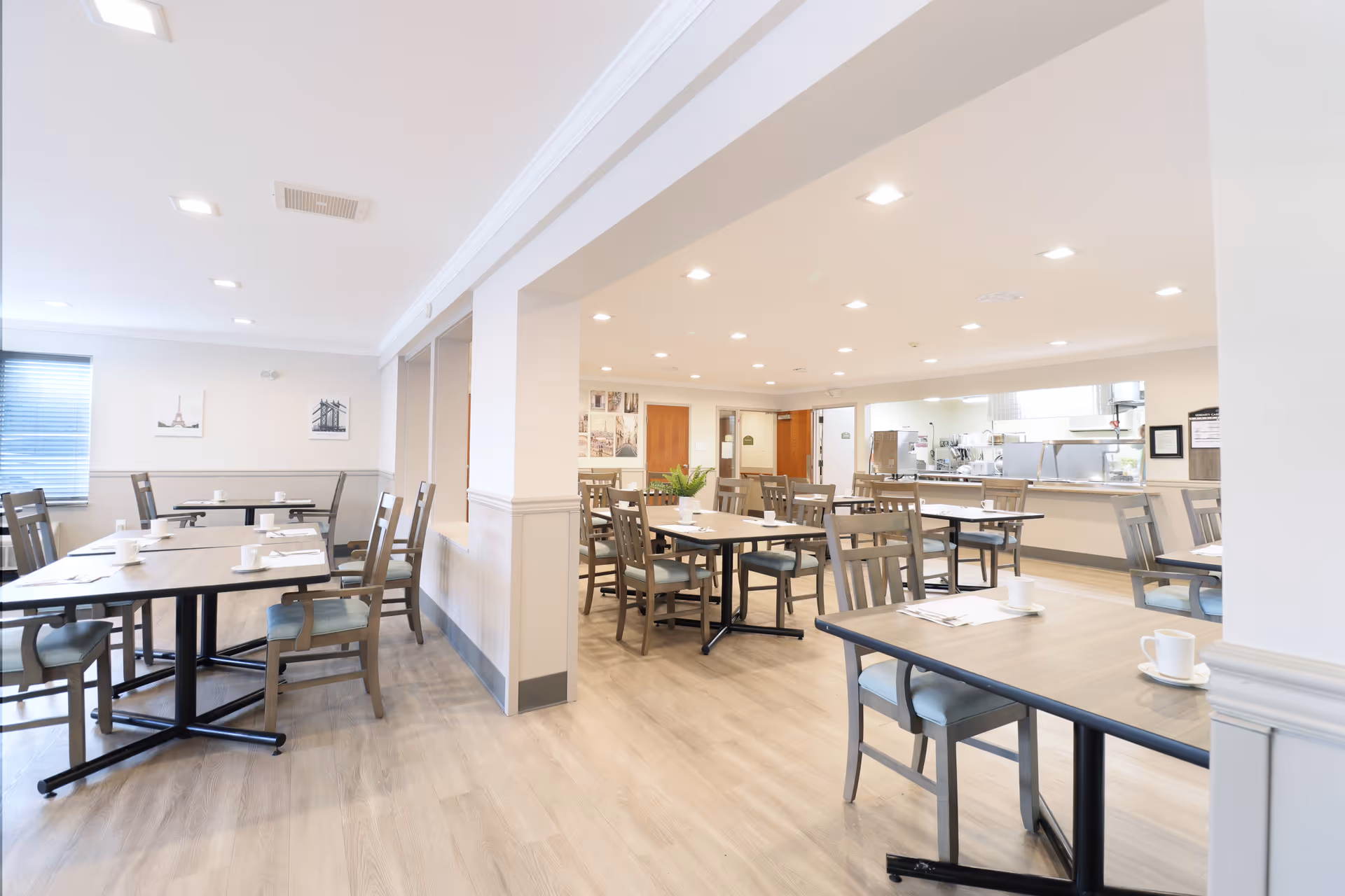 A bright and clean dining room with multiple wooden tables and chairs arranged neatly. Each table has white cups and napkins placed on it. The room has light-colored walls, wood flooring, and recessed lighting in the ceiling. There are framed pictures on the walls and a window with blinds on the left side. In the background, a kitchen area with stainless steel appliances and counters is visible.