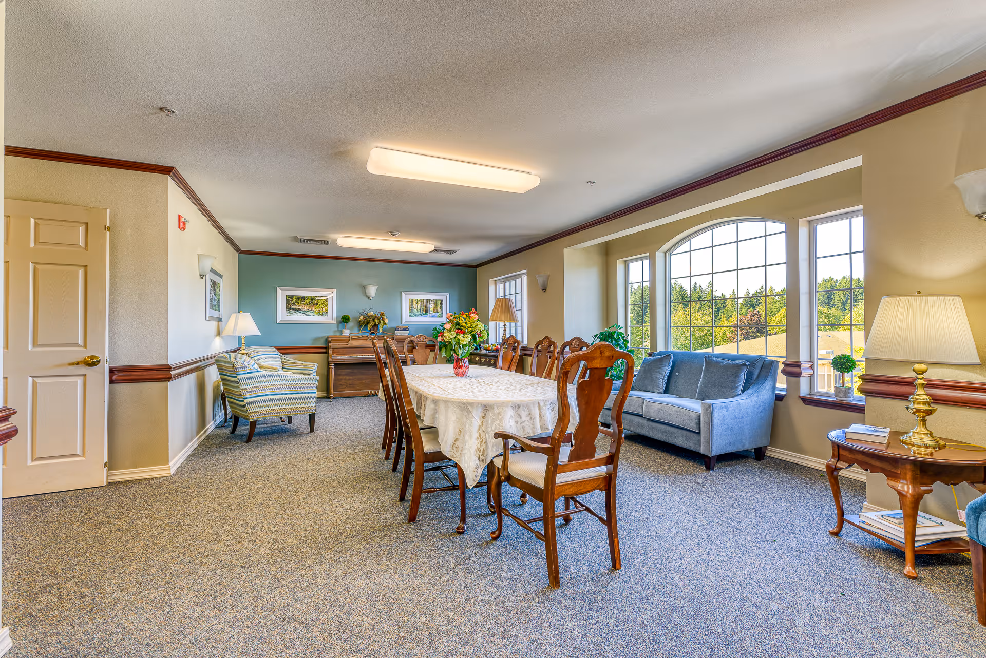 A bright and spacious common area in an assisted living facility featuring a long dining table with a white tablecloth and wooden chairs, a blue sofa near large windows with a scenic outdoor view, a small side table with a lamp and books, and a piano against a teal accent wall with framed pictures and decorative plants.