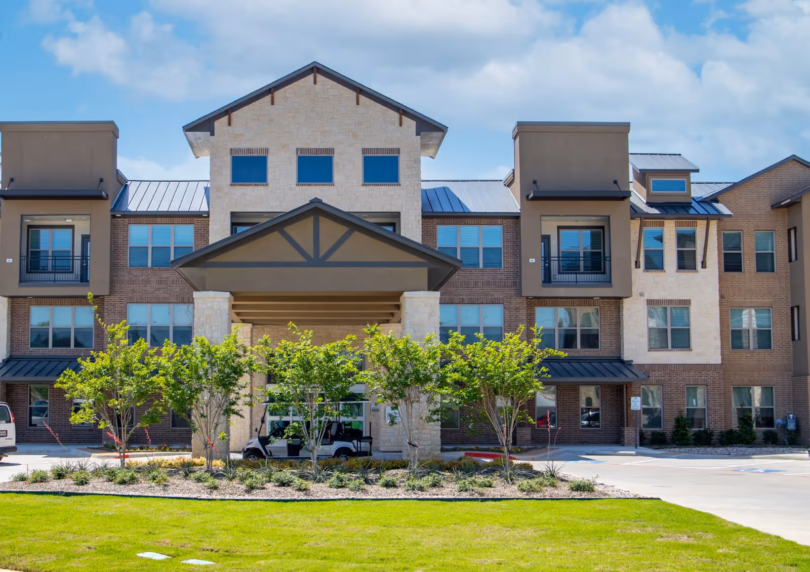 Front exterior view of a multi-story senior living facility building with a covered entrance, stone and brick facade, multiple windows, and landscaped greenery in front under a partly cloudy sky.