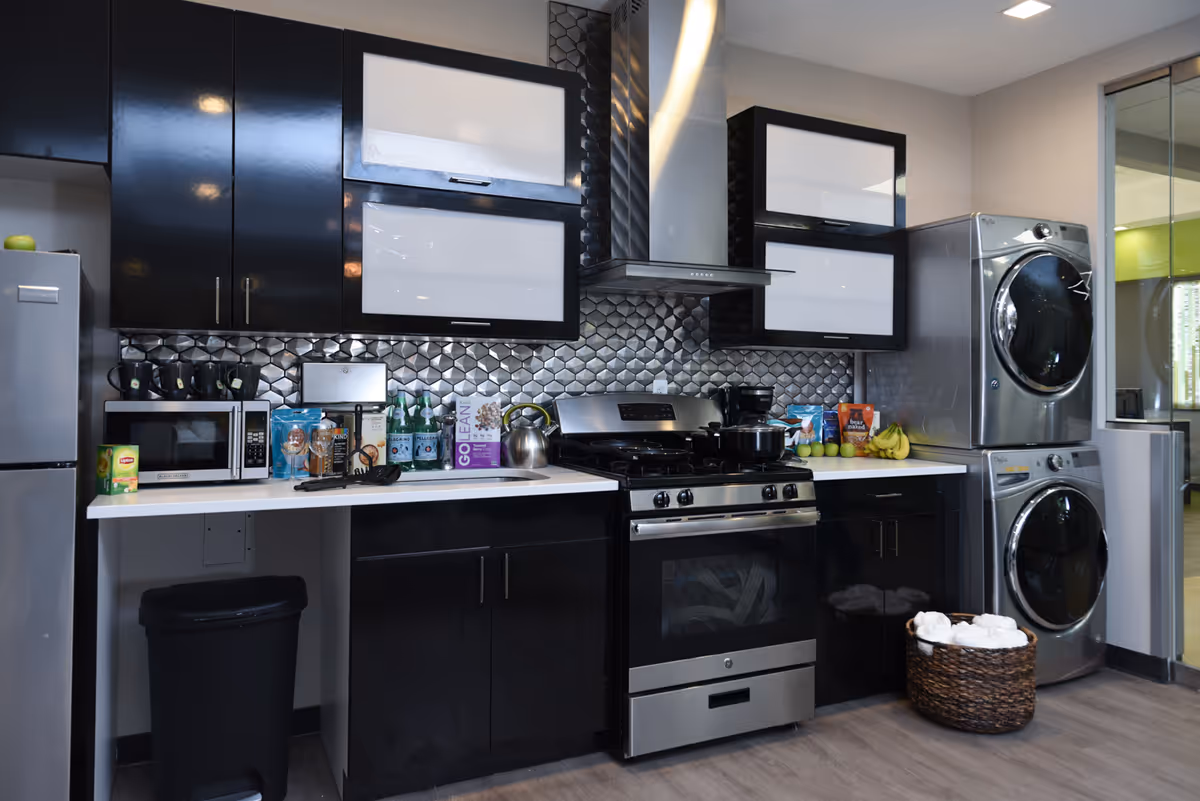 Modern kitchen area with black and white cabinets, a stainless steel stove with a range hood, a microwave, a refrigerator, and a stacked washer and dryer. The countertop has various items including bottles, snacks, and a kettle. A woven basket with towels is placed on the floor next to the washer and dryer.