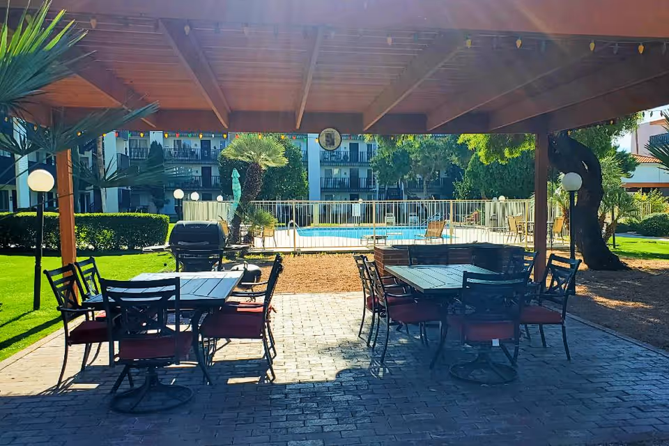 Outdoor covered patio area with two tables and several chairs with red cushions, a barbecue grill, and a view of a fenced swimming pool surrounded by greenery and apartment balconies in the background.