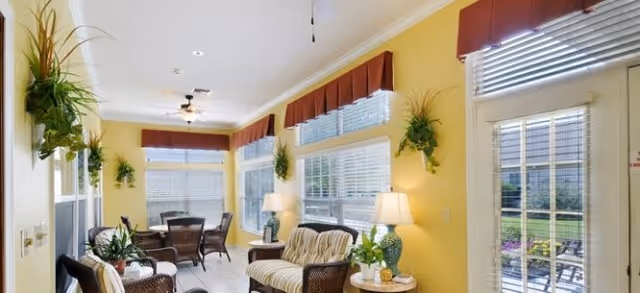 A bright sunroom with yellow walls and multiple windows covered with white blinds and red valances. The room is furnished with wicker chairs and a loveseat with cushions, a round dining table with chairs, several potted plants, and two table lamps. A ceiling fan is mounted on the white ceiling, and a glass door leads outside to a garden area.