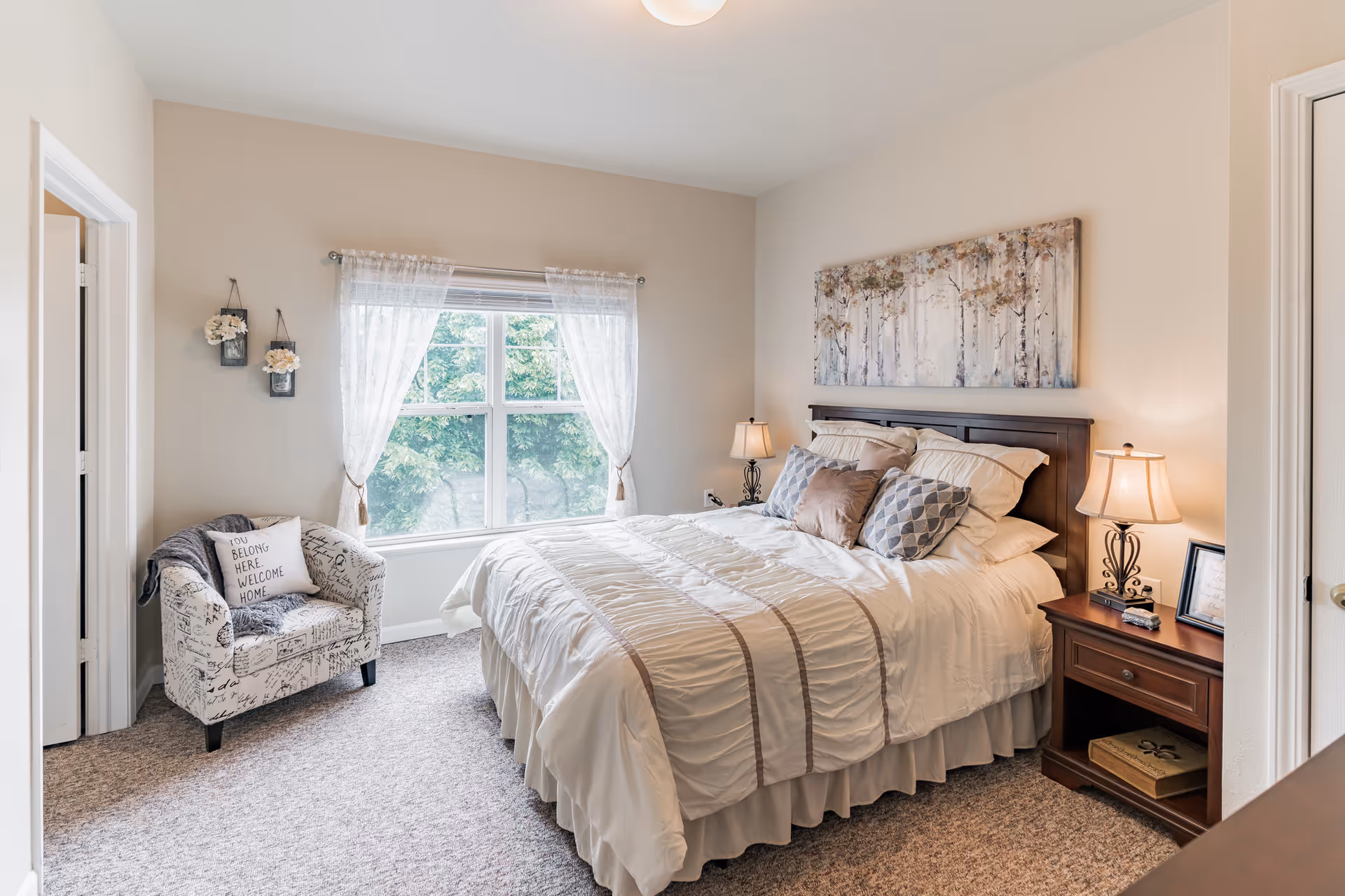 A cozy bedroom with a large bed covered in white and beige bedding, two bedside tables with lamps, a decorative painting above the bed, a window with sheer curtains letting in natural light, and a patterned armchair with a pillow that reads 'You belong here. Welcome home.'