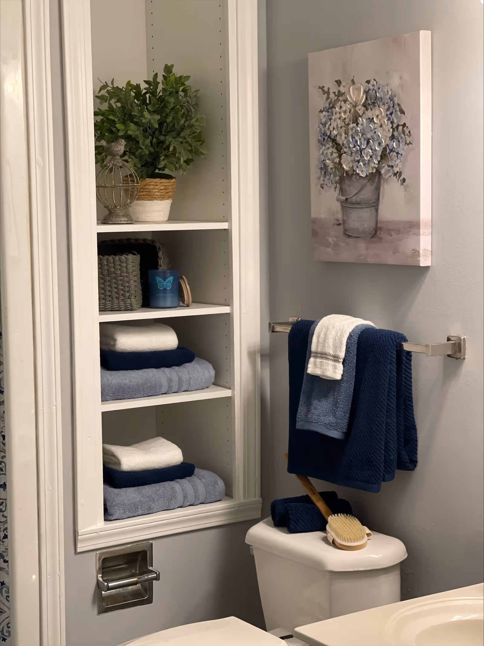 A bathroom corner featuring a built-in white shelving unit with neatly folded towels, a small plant, a decorative birdcage, and a blue candle. Next to the shelves is a towel rack with blue and white towels hanging above a toilet. A wooden bath brush rests on the toilet tank. A painting of a bucket with blue and white flowers hangs on the wall above the towel rack.