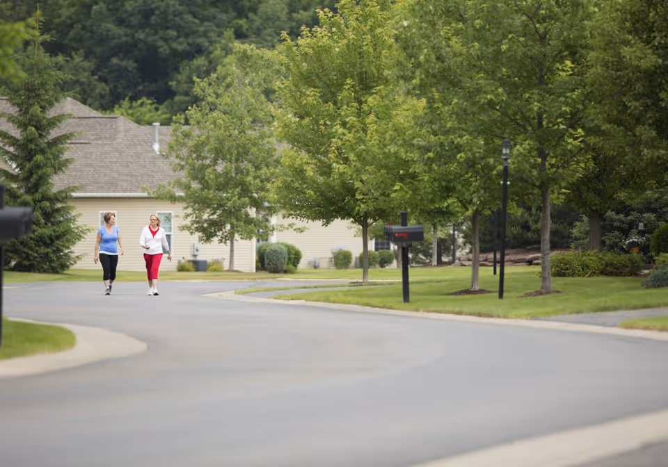 Two women walking along a tree-lined street in a residential neighborhood of townhomes with mailboxes and lawns.