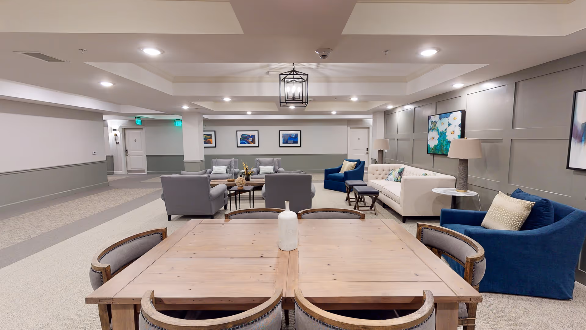 A spacious and well-lit common living area in a senior living facility featuring a wooden table with six chairs in the foreground, a white vase centerpiece, multiple comfortable armchairs and sofas in shades of gray, blue, and beige, decorative pillows, two table lamps on side tables, framed artwork on the walls, and recessed ceiling lights.