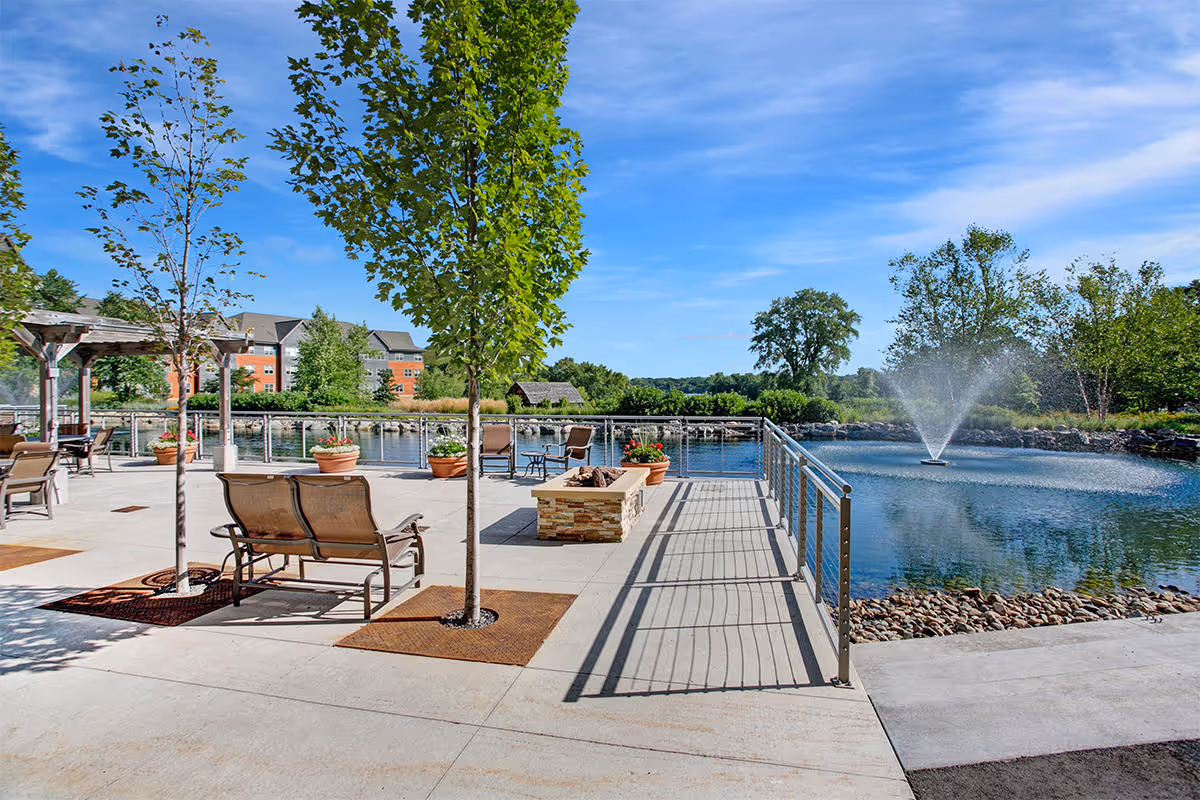 Outdoor patio area with seating benches, potted plants, young trees, and a fire pit. There is a metal railing overlooking a pond with a water fountain in the center. In the background, there are trees, a building, and a clear blue sky.