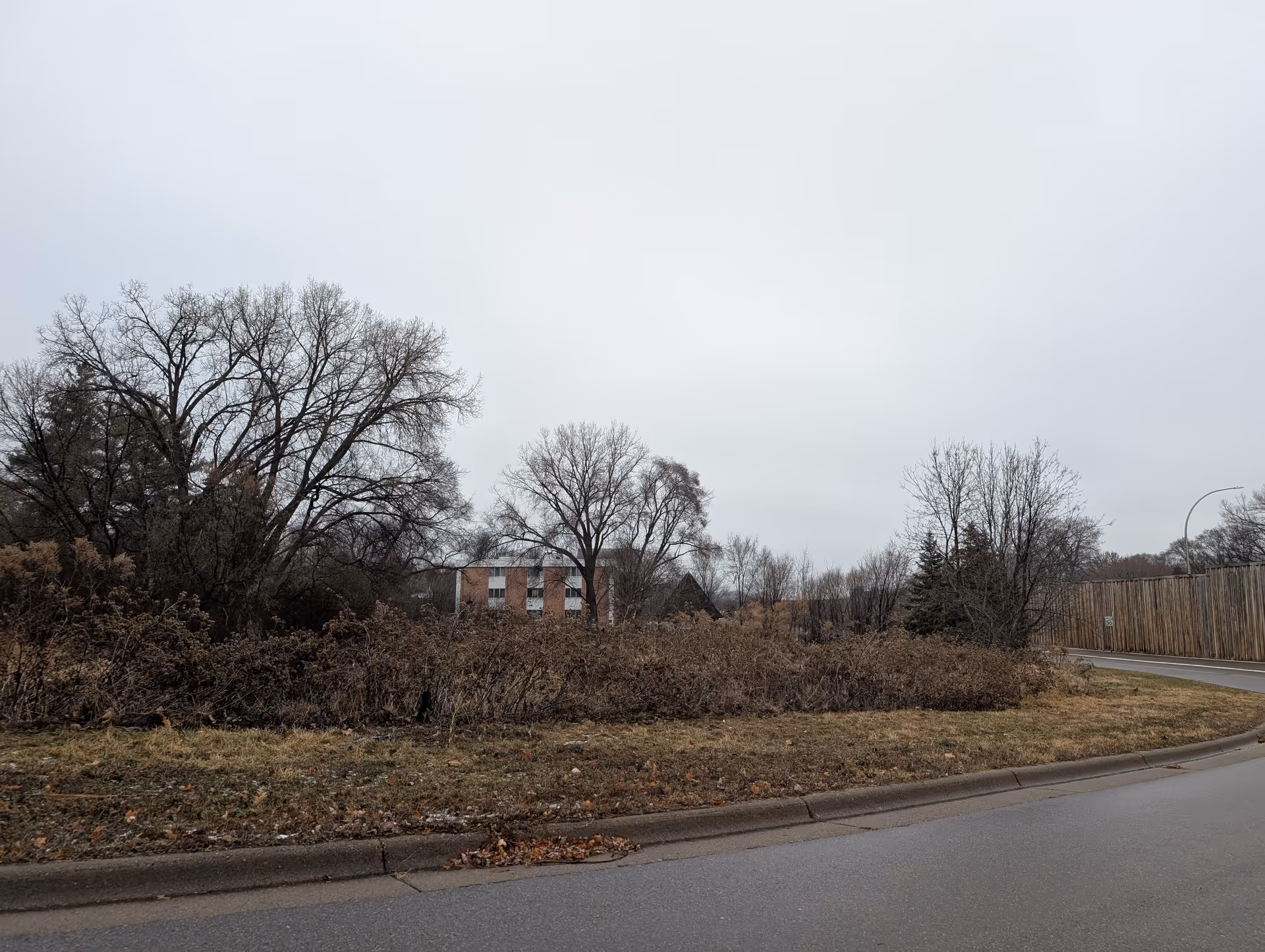 View of a road curving to the right with a speed limit sign, bordered by dry grass and bushes. Behind the bushes are leafless trees and a partially visible brick building under an overcast sky.