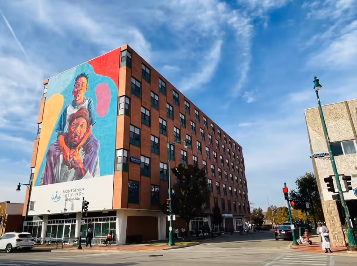 Street view of a multi-story senior living facility building with a large colorful mural of two people on the side wall. The building has many windows and is located at a street intersection with traffic lights and pedestrians.