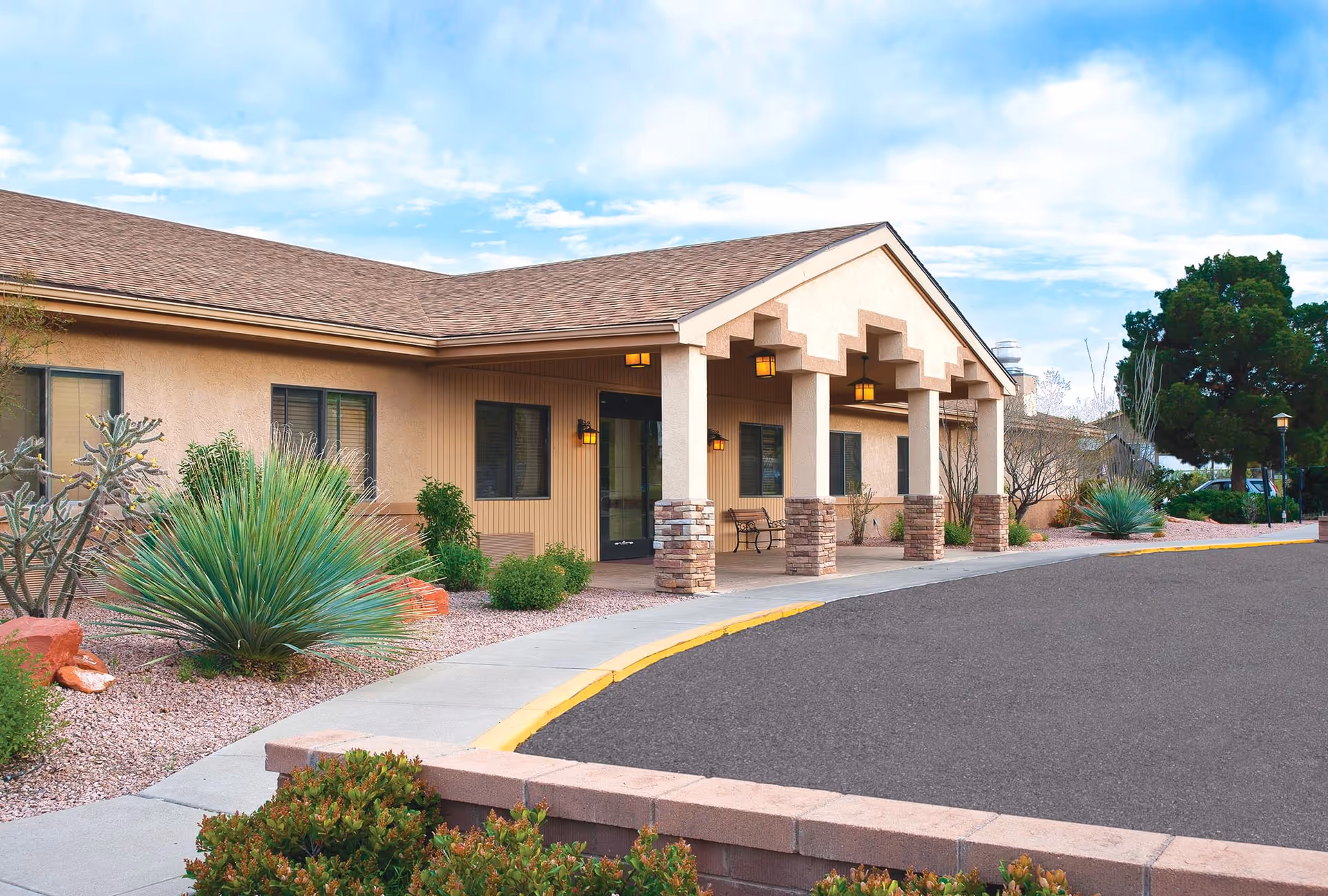 Exterior view of a single-story building with a covered entrance supported by columns with stone bases. The building is surrounded by desert landscaping with various shrubs and cacti, and a paved driveway curves in front of the entrance under a partly cloudy sky.