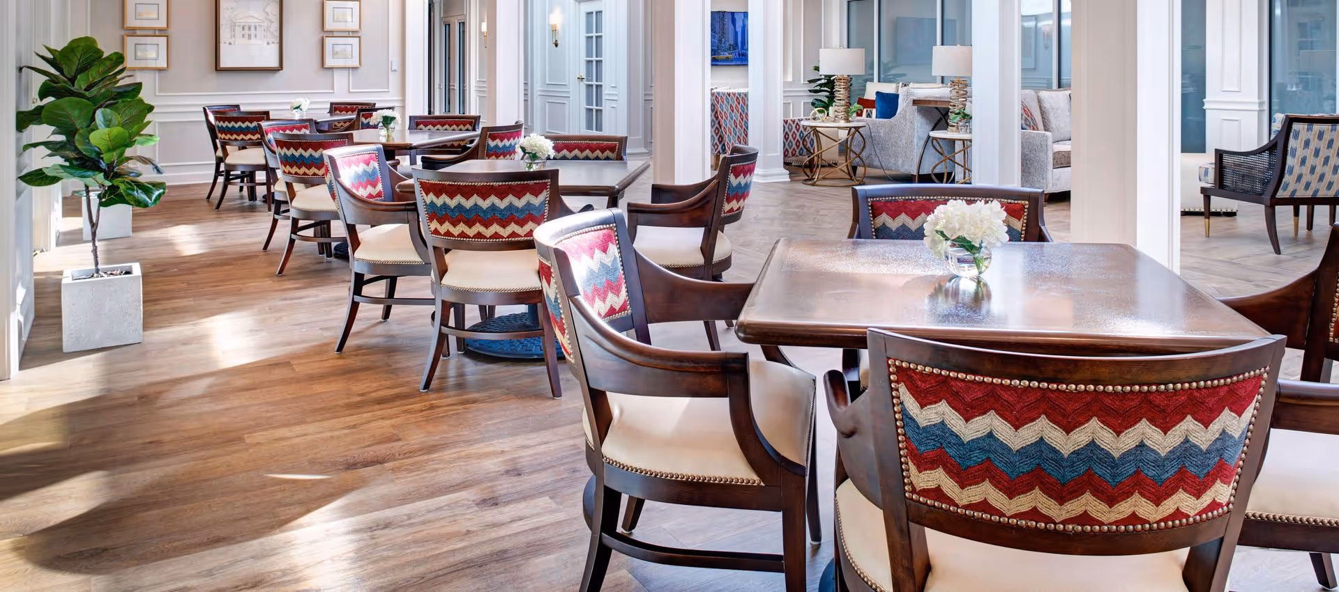 A bright and elegant dining area with wooden tables and chairs featuring colorful zigzag patterned upholstery. The room has wood flooring, white walls with decorative molding, framed artwork, and potted plants. In the background, there is a cozy seating area with sofas and side tables with lamps.