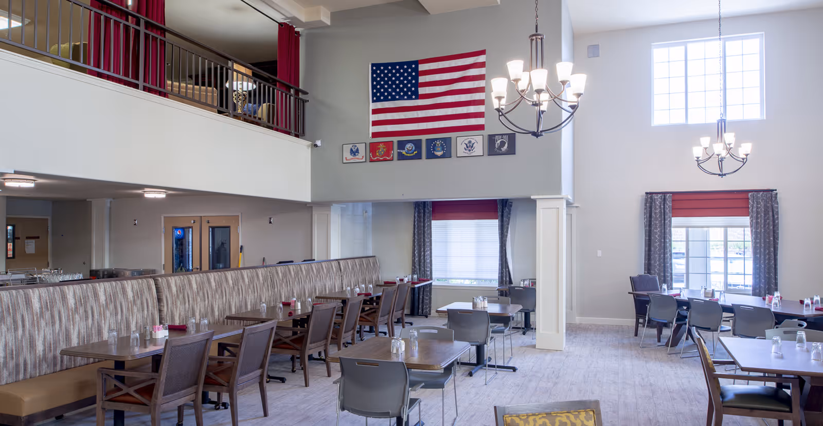A spacious senior living dining room with multiple tables and chairs arranged neatly. The room features high ceilings, large windows with red and patterned curtains, and two chandeliers hanging from the ceiling. An American flag and military service flags are displayed on the wall above a half wall with a railing overlooking the dining area.