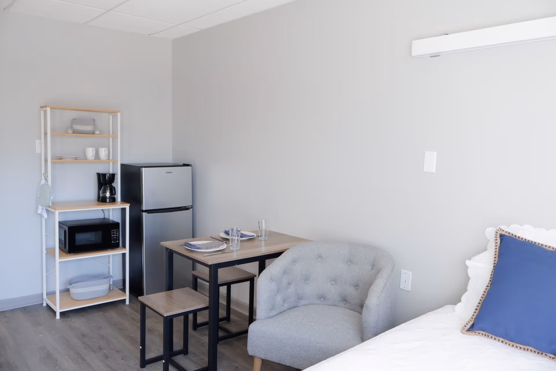 A small living space with a light gray armchair, a wooden table set with two plates and glasses, a small refrigerator, a microwave on a white and wood shelving unit, and part of a bed with a blue pillow.