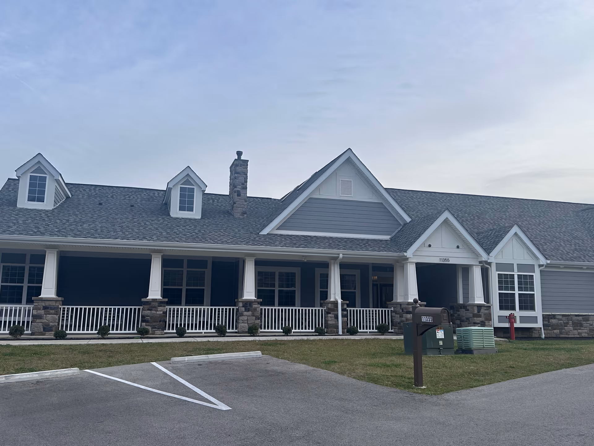 Exterior view of a single-story building with a gray roof, white trim, and stone accents. The building has a covered porch with white railings and columns. There is a mailbox and some utility boxes on the lawn in front of the building, along with a parking area.