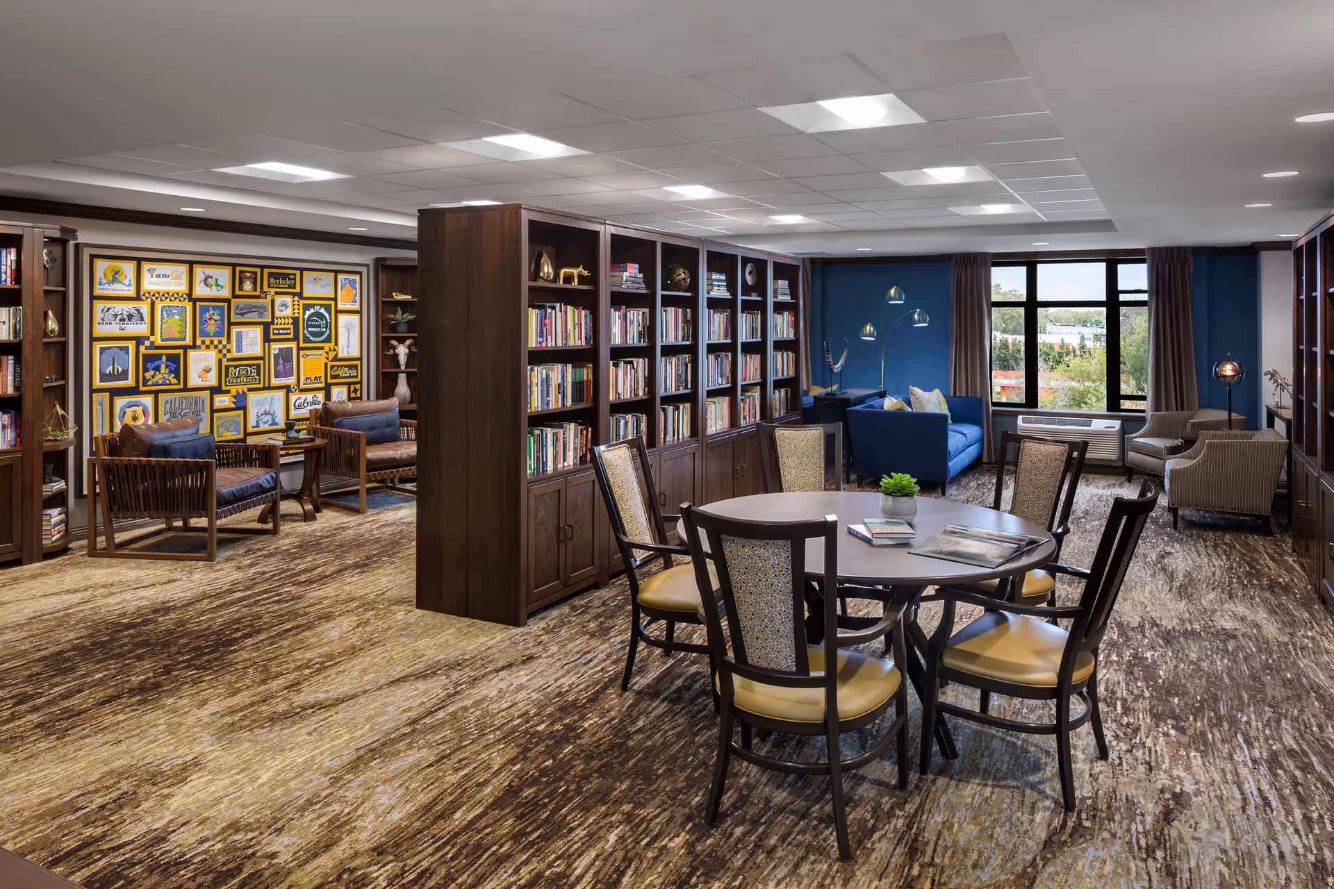 A spacious senior living community library/lounge with bookshelves, seating areas, a round table with chairs, and large windows.