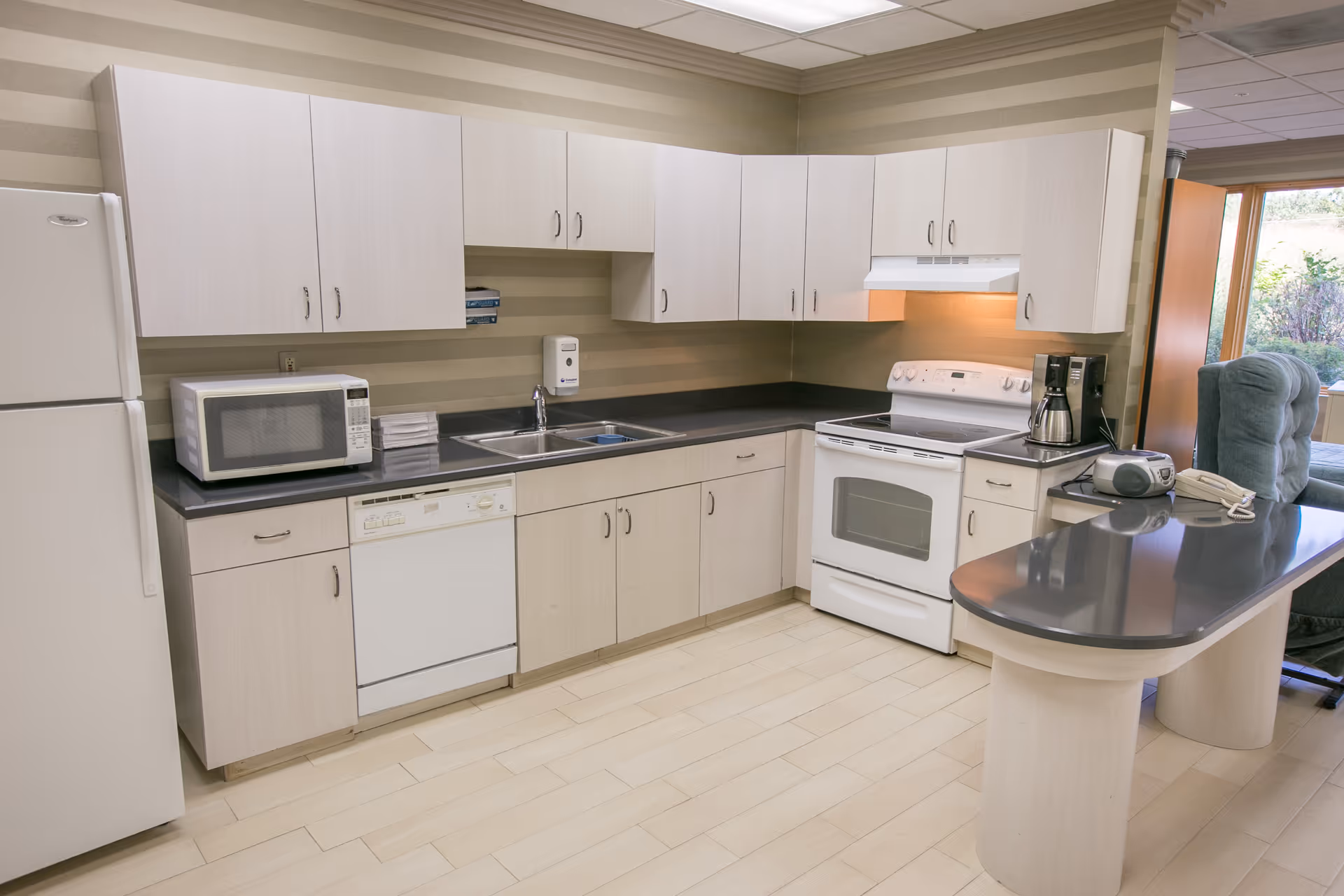 A clean and modern kitchen area with light wood cabinets, a white refrigerator, microwave, dishwasher, stove with oven, coffee maker, and a countertop with a telephone and radio. The kitchen has a striped beige wall and light-colored flooring, with a window showing some greenery outside.