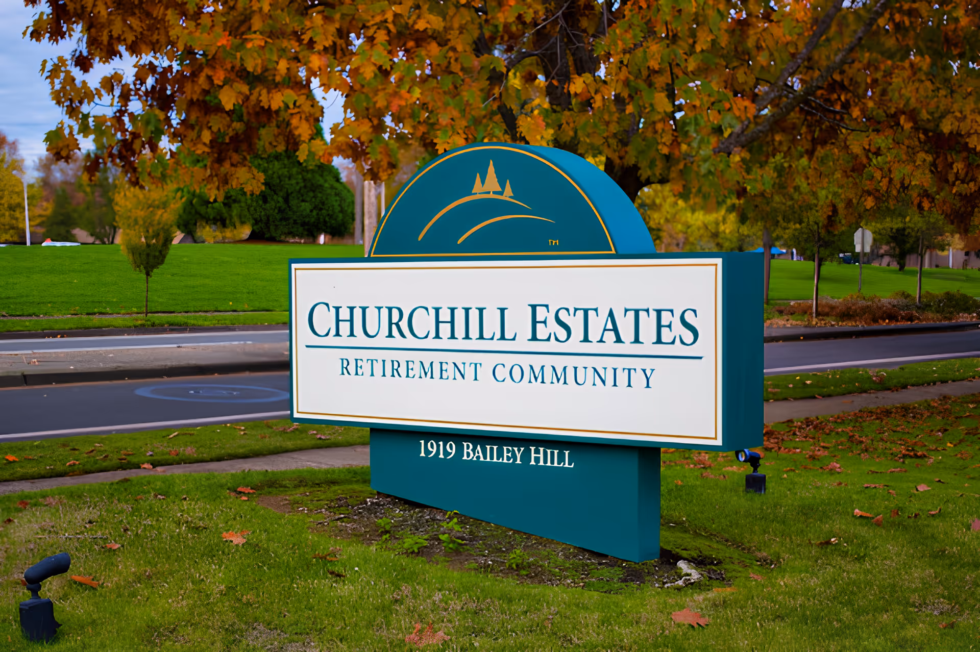 A green and white entrance sign reading "Churchill Estates Retirement Community" on a lawn beneath autumn trees.