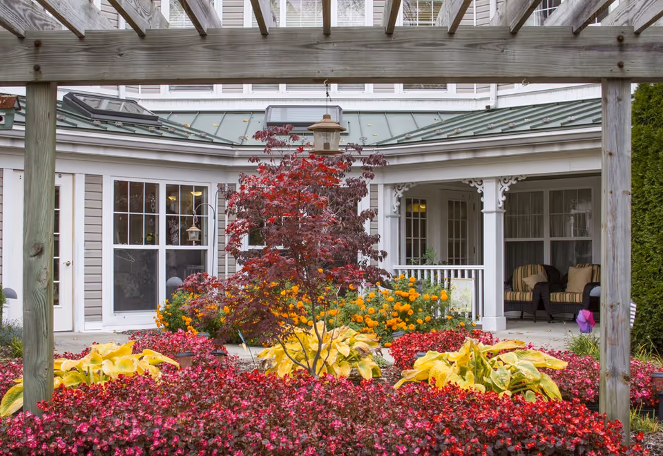 A landscaped courtyard with red and yellow flowers and a small ornamental tree framed by a wooden pergola in front of a building porch.