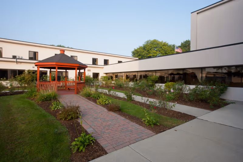 Outdoor courtyard with a red wooden gazebo, brick walkway, planted garden beds, and a surrounding two-story building.