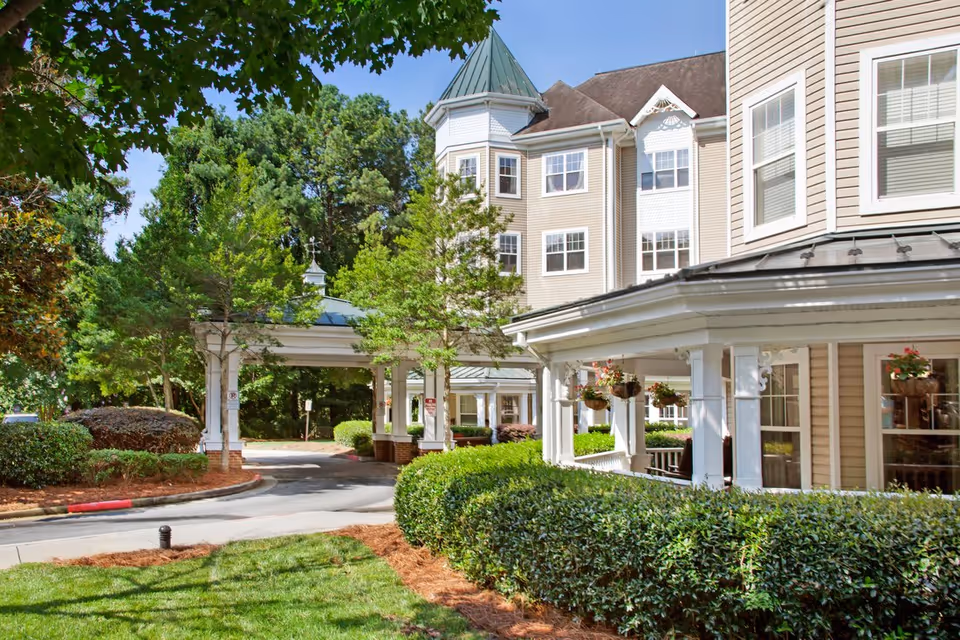 Exterior view of a senior living facility building with beige siding, white trim, and multiple windows. The building features a covered entrance with white columns and hanging flower baskets. Surrounding the building are green bushes, trees, and a well-maintained lawn under a clear blue sky.