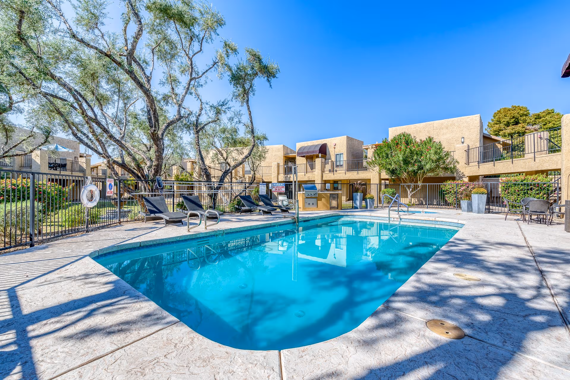 Outdoor swimming pool area at Scottsdale Village Square with lounge chairs, a barbecue grill, and surrounding two-story beige buildings under a clear blue sky.