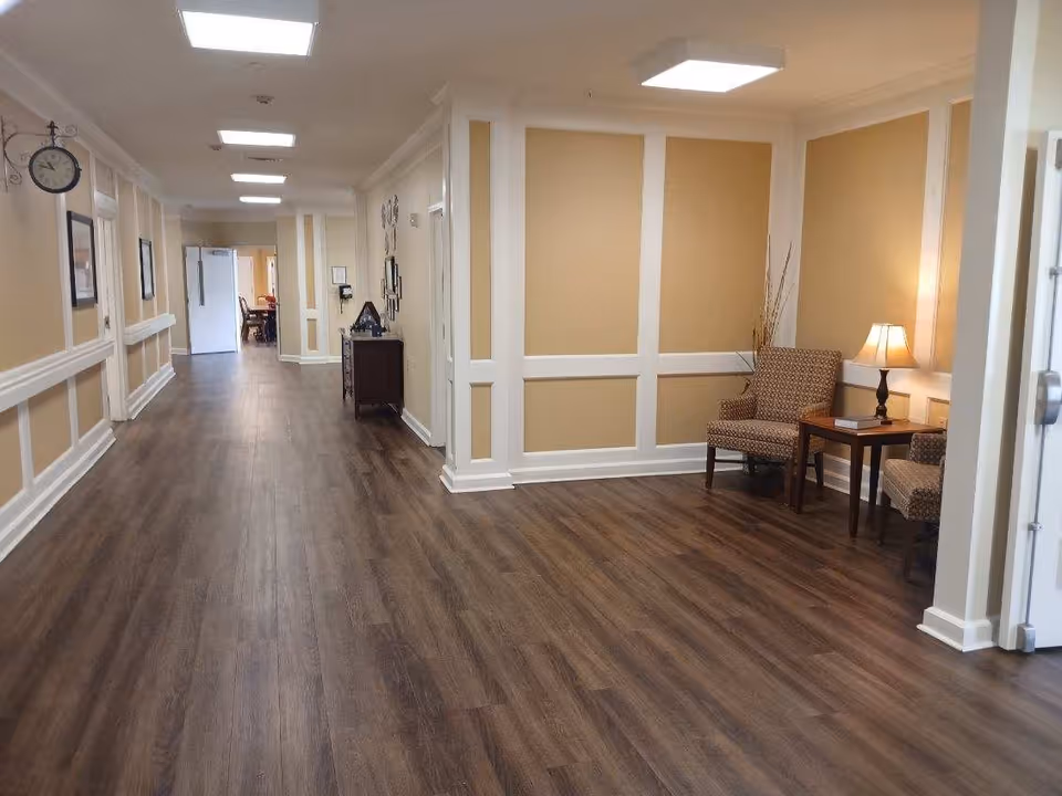 A long hallway with wood flooring and beige walls with white trim. On the right side, there is a small seating area with two patterned armchairs, a wooden side table with a lamp, and decorative dried plants. The hallway is well-lit with ceiling lights and has a clock and framed pictures on the left wall. At the far end, a dining area with tables and chairs is visible.
