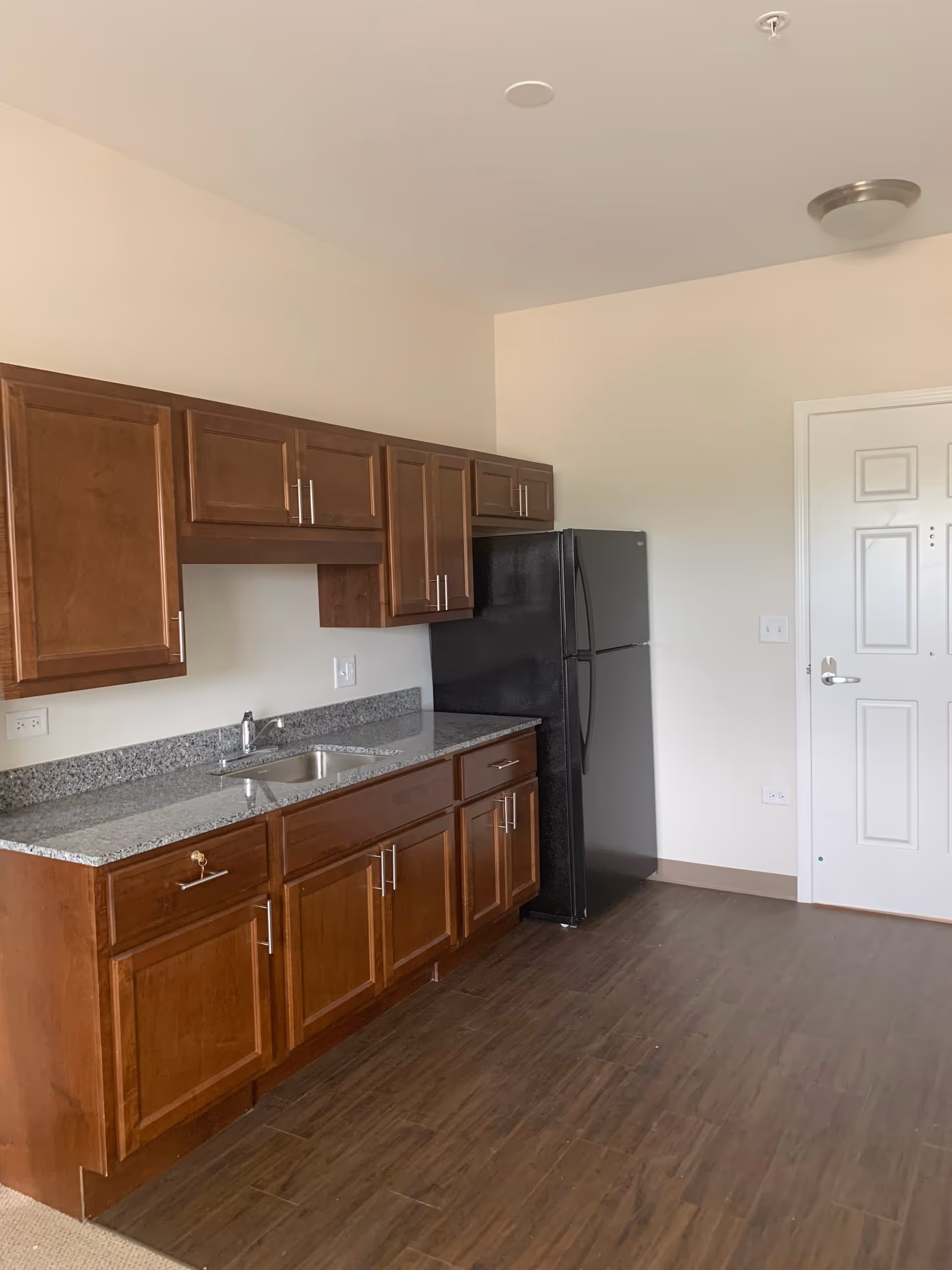 Interior view of a kitchen area with wooden cabinets, a granite countertop with a sink, a black refrigerator, and a white door. The floor is wood-style, and the walls are painted light beige.