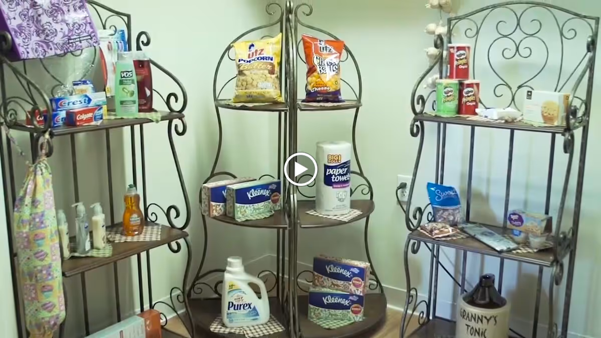 Three black metal corner shelves arranged in a room corner, stocked with various household items including snacks like popcorn and chips, cleaning supplies such as paper towels and laundry detergent, personal care products like toothpaste and lotion, and other miscellaneous items.