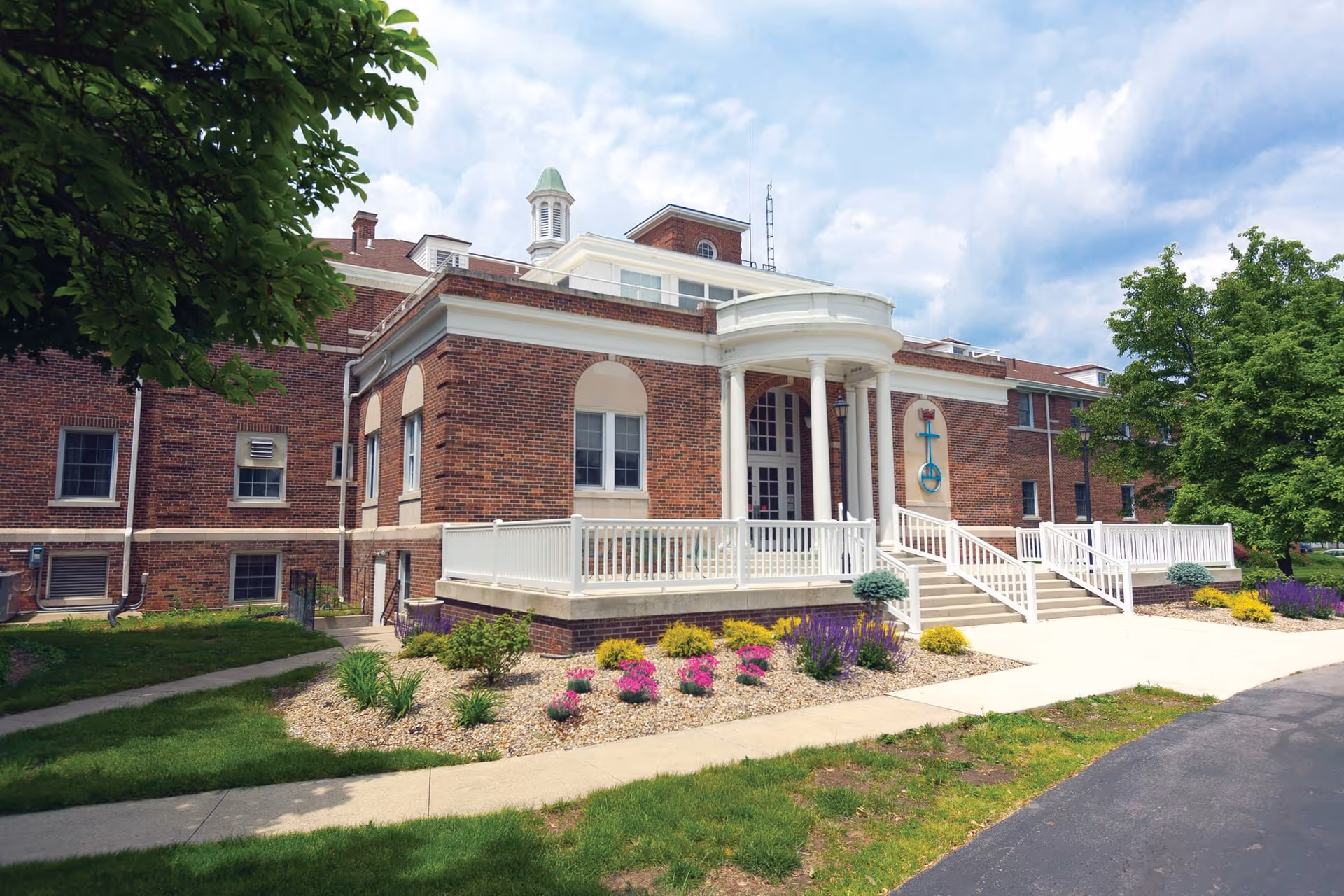 Exterior view of a brick senior living facility building with white columns and railings at the entrance. There are landscaped flower beds with colorful flowers and green shrubs along the walkway leading to the entrance. Trees and a partly cloudy sky are visible in the background.