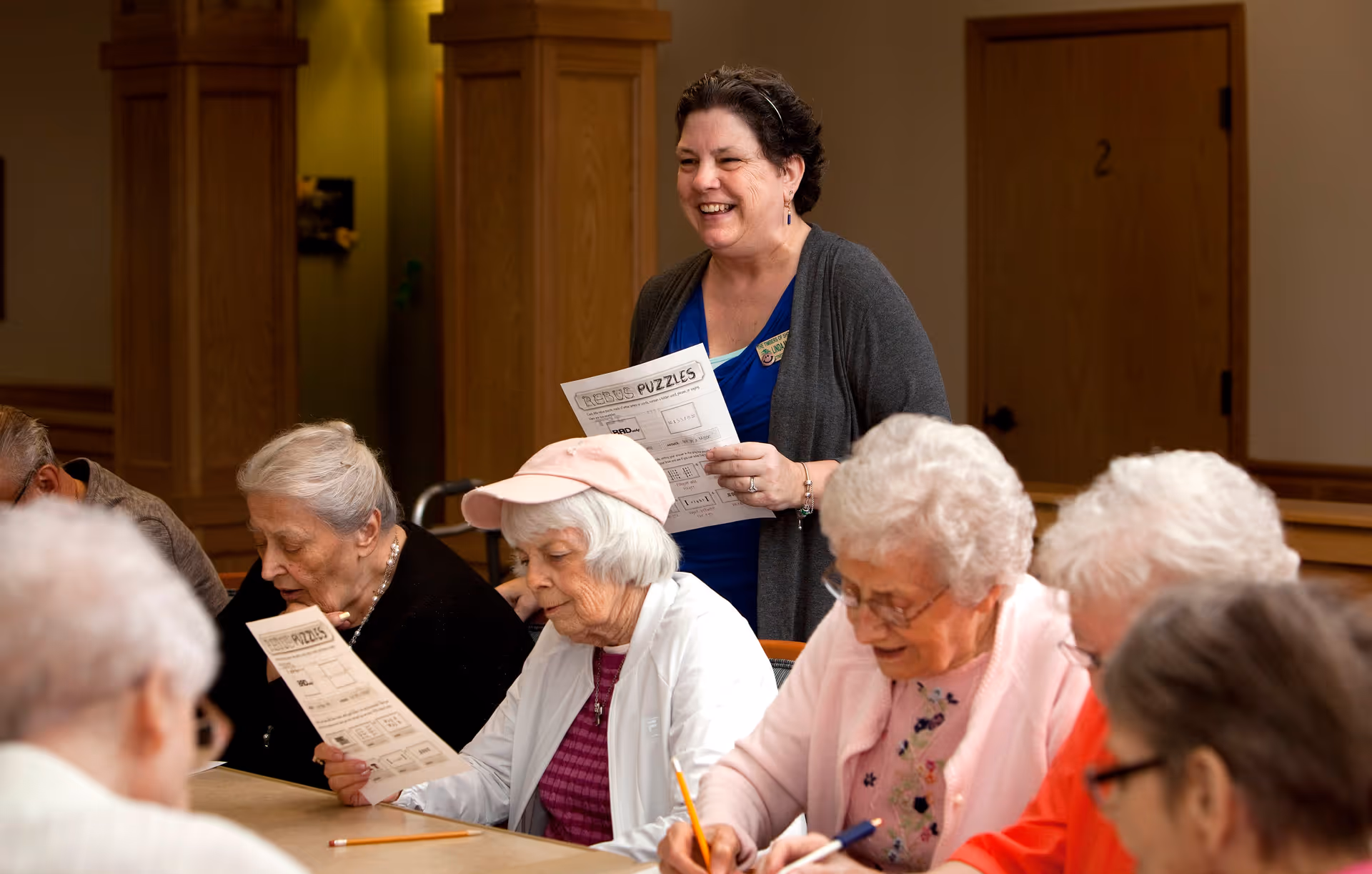 A group of elderly women sitting around a table working on puzzles or worksheets, while a smiling woman stands behind them holding a puzzle sheet, in a warm, wood-paneled room.